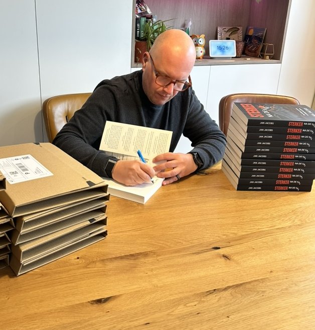 Jan signeert een exemplaar aan tafel, met de verzenddozen ernaast